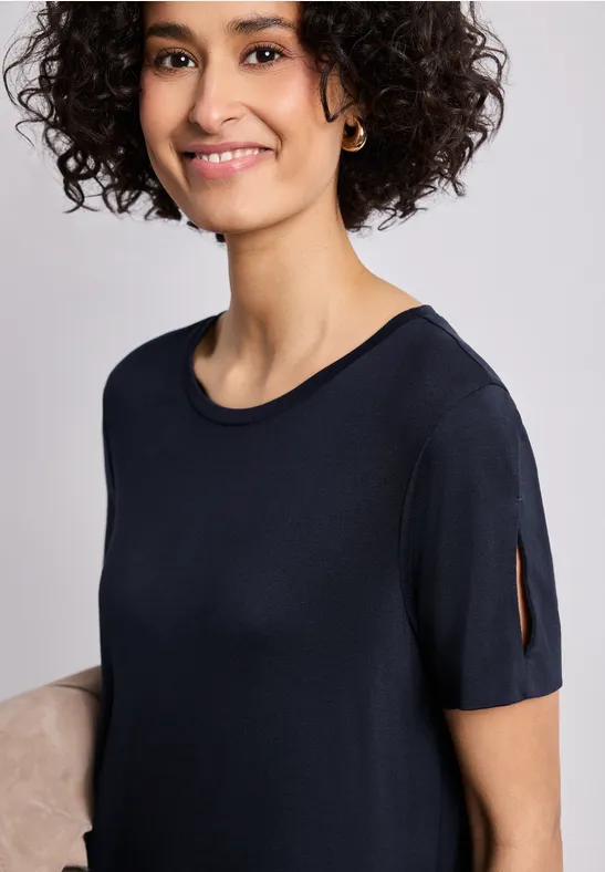 Femme aux cheveux bouclés et créoles porte un t-shirt noir à manches courtes fendues, visage flou.