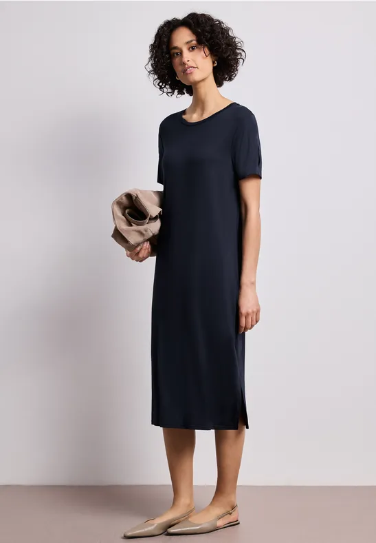 Femme porte une robe midi bleu marine à manches courtes et des ballerines beige, tenant un gilet clair.
