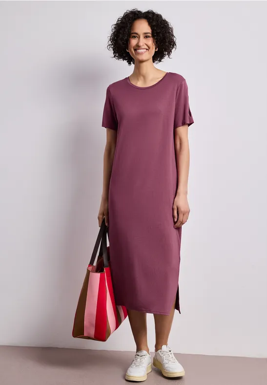 Persoon in paarse midi T-shirtjurk, witte sneakers en een grote kleurrijke draagtas.