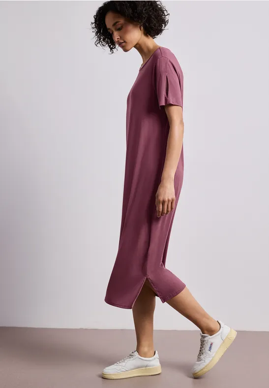 Zijwaarts lopend persoon in losvallende midi-jurk roze-paars en witte sneakers.