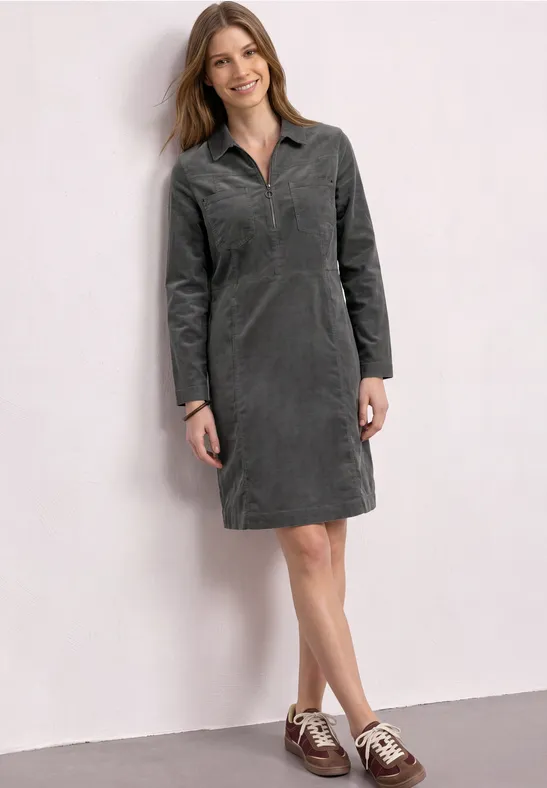 Femme en robe-chemise en velours côtelé gris, manches longues et col, avec derbies bicolores devant un mur blanc.