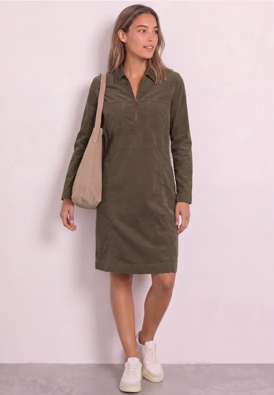 Frau in olivgrünem Cordkleid mit Kragen und langen Ärmeln, weißer Sneaker und beige Tasche.