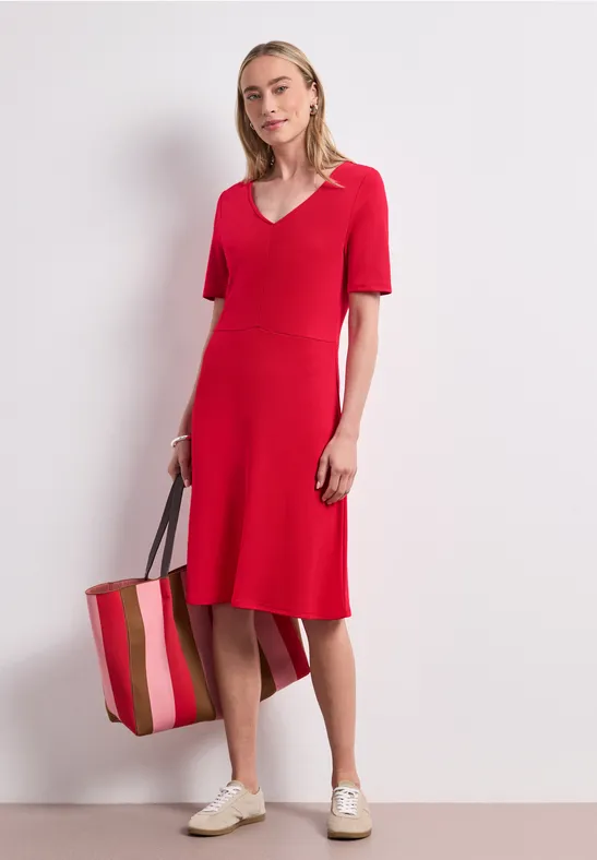 Vrouw in halflange rode jurk met V-hals en korte mouwen, met gestreepte tas en witte sneakers.