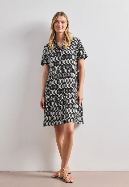 Femme portant une robe mi-longue à motifs noir et blanc, manches courtes et sandales, devant un mur clair.