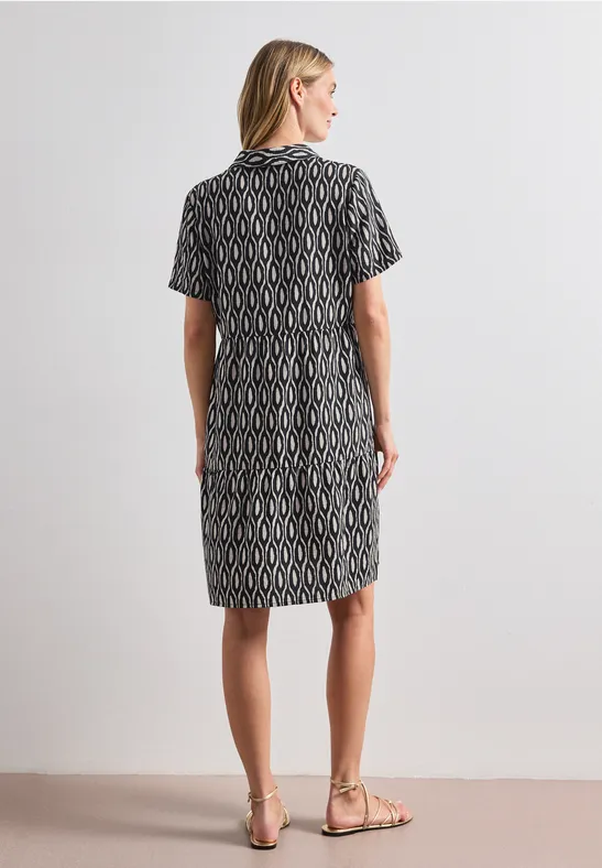 Femme vue de dos, en robe noire à motifs jusqu'aux genoux et sandales, sur fond neutre.