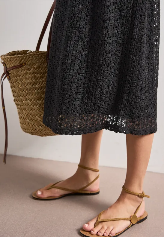 Vrouw in zwarte gehaakte jurk, rieten tas en platte, olijfkleurige sandalen met bandjes.