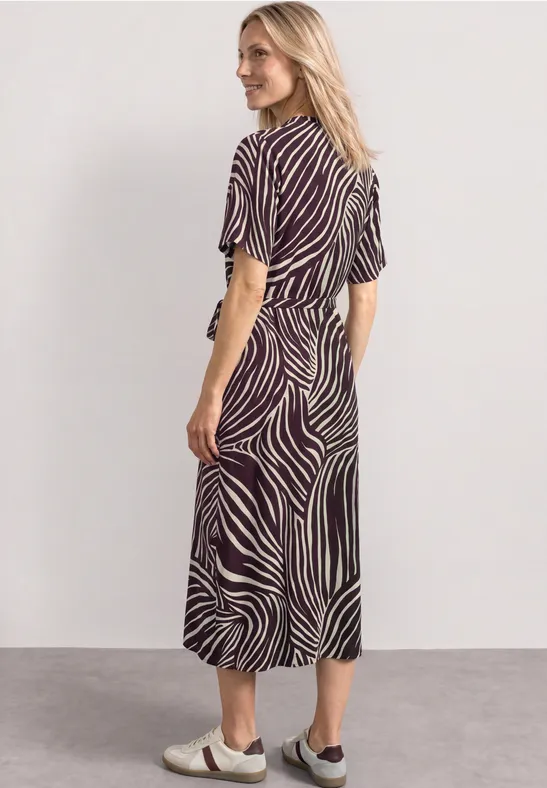 Een vrouw in een blauw-witte zebraprint midi-jurk met witte slippers staat zijwaarts voor een grijze achtergrond.