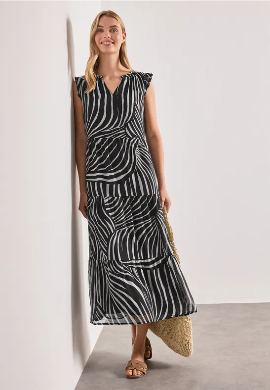 Vrouw leunend tegen muur in elegante zwart-witte zebradress met ceintuur en strooien tas.