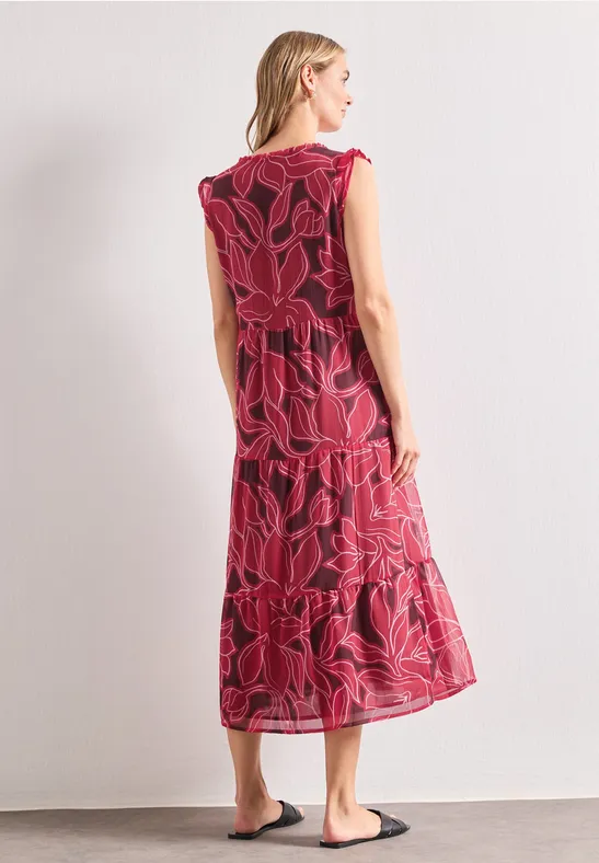 Vrouw van achteren in een rood midilange jurk met groot bloemenpatroon en zwarte sandalen tegen lichte achtergrond.
