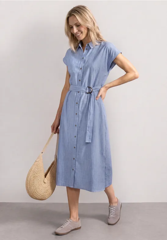 Frau in hellblauem Hemdkleid mit Bindegürtel, Sneakers und Strohtasche.