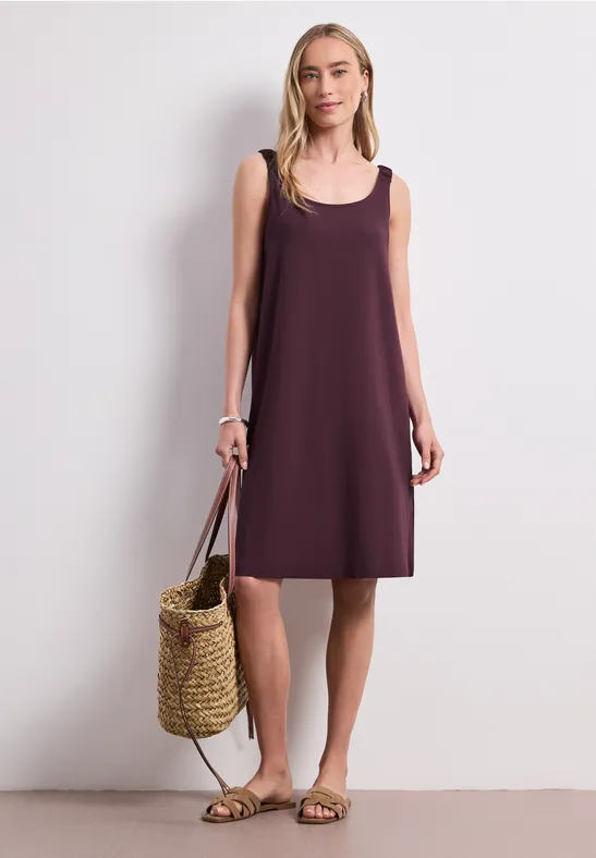 Vrouw in losse mouwloze paarse jurk met een strooien tas en sandalen.