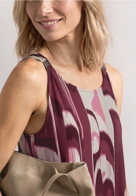 Vrouw in mouwloze bordeauxrode, geprinte jurk met beige schoudertas.