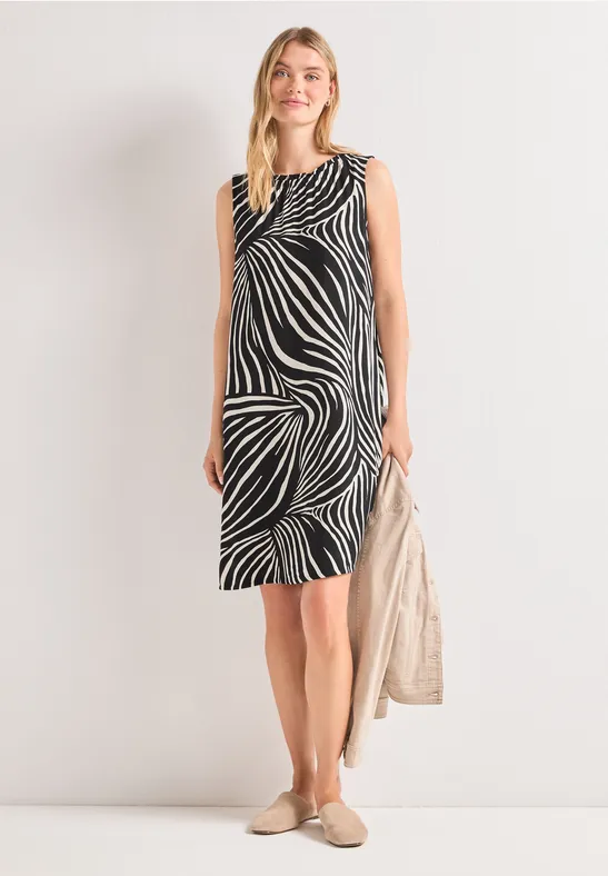 Vrouw in mouwloze, knielange jurk met zwart-wit zebraprint, houdt een lichte tas vast.
