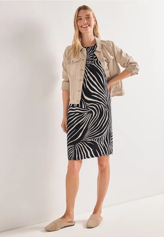 Vrouw in knielange jurk met zwart-witte zebraprint, beige jasje en comfortabele instappers.