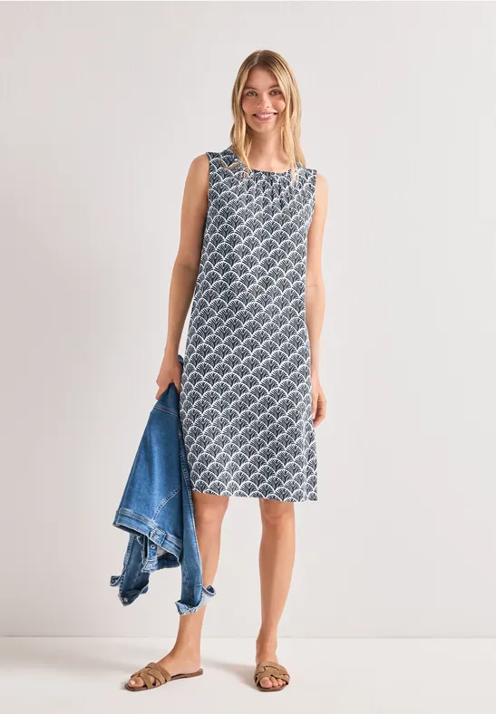 Vrouw in mouwloze, knielange jurk met dessin, sandalen aan en een spijkerjasje in de hand.