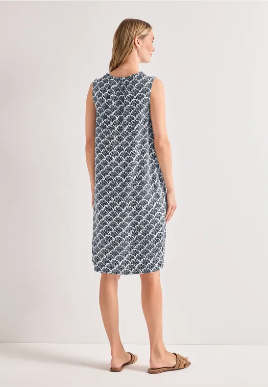 Vrouw van achteren in een mouwloze, halflange blauw-witte jurk met dessin en platte sandalen.