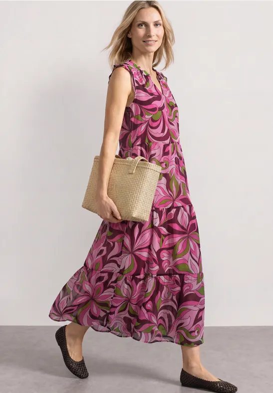 Vrouw in roze-groene bloemenjurk, rieten tas en zwarte ballerina’s lopend op straat.