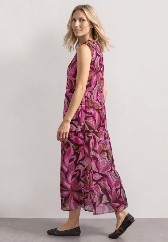 Vrouw in zijaanzicht draagt een lange mouwloze maxi-jurk met roze-paars bloemenpatroon en platte schoenen.