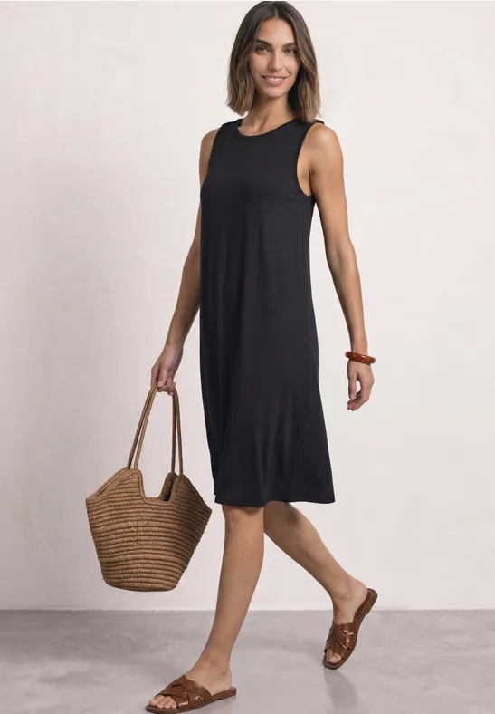 Frau in ärmellosem schwarzen Midi-Kleid, mit Strohtasche und braunen Sandalen.