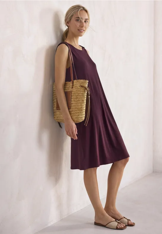 Frau in ärmellosem violettem Kleid, trägt geflochtene Korbtasche und Sandalen, lehnt an heller Wand.