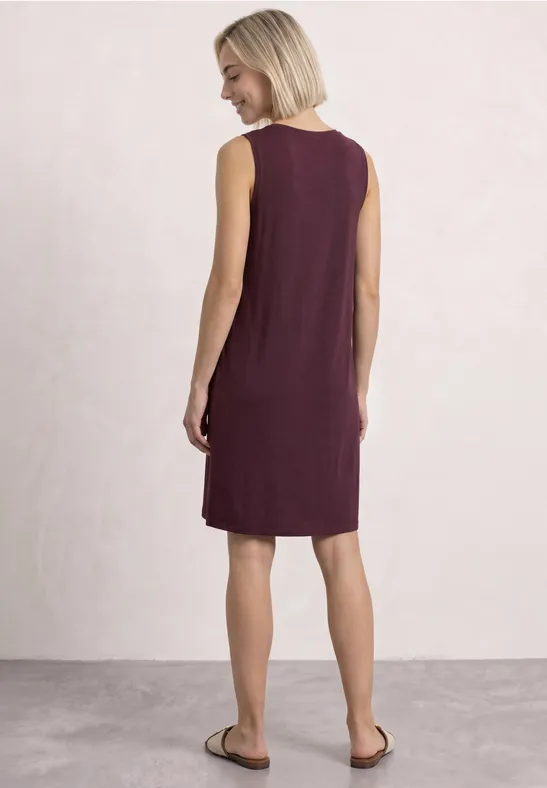 Frau von hinten in ärmellosem, knieumspielendem, dunkelrotem Kleid und Sandalen vor hellem Hintergrund.