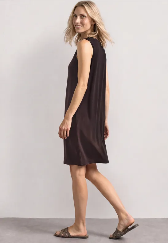 Femme en robe noire sans manches et sandales marchant de profil sur fond clair.