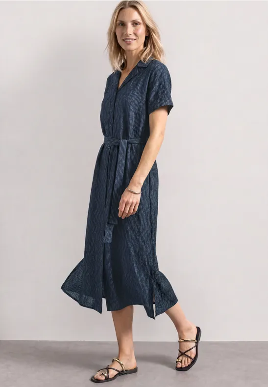 Vrouw in blauwe midi-jurk met ceintuur en zwarte sandalen.
