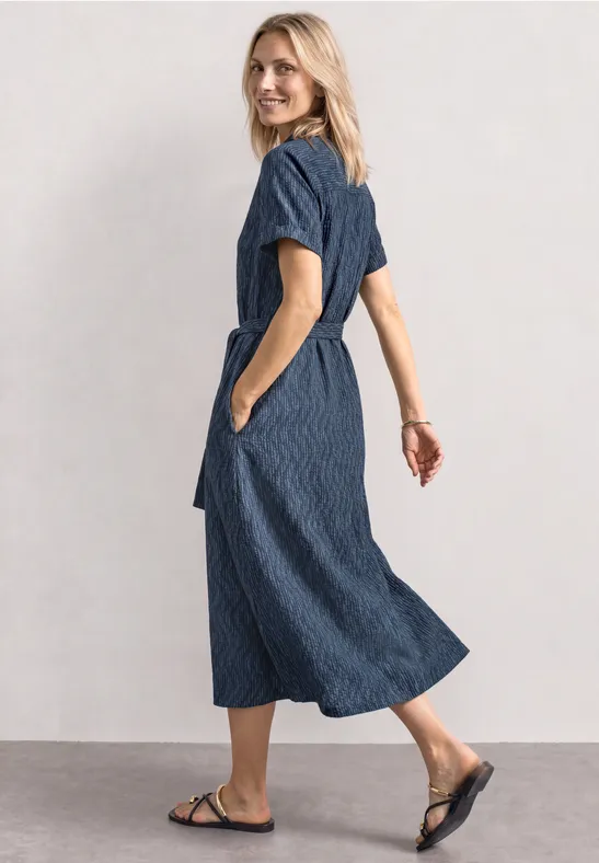 Vrouw in blauwe, midilengte jurk met zakken en zwarte sandalen tegen een neutrale achtergrond.