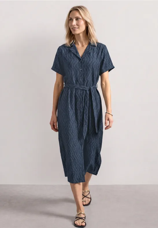 Vrouw in getailleerde navyblauwe midi-jurk met korte mouwen, V-hals en ceintuur tegen neutrale achtergrond