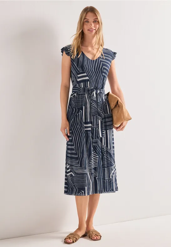 Vrouw in blauw-witte midi jurk met ceintuur, sandalen en beige clutch.
