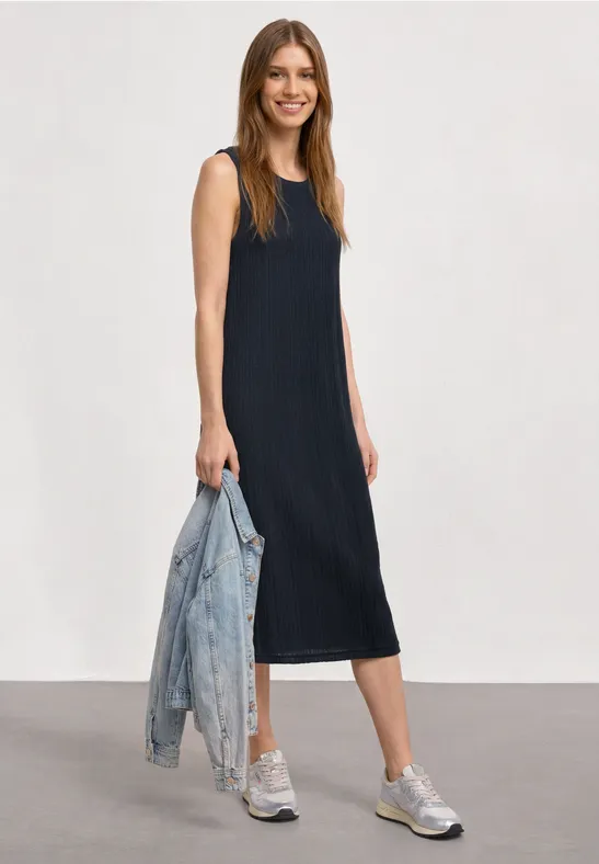 Vrouw in mouwloze midi-jurk marineblauw, witte sneakers en een spijkerjasje in de hand