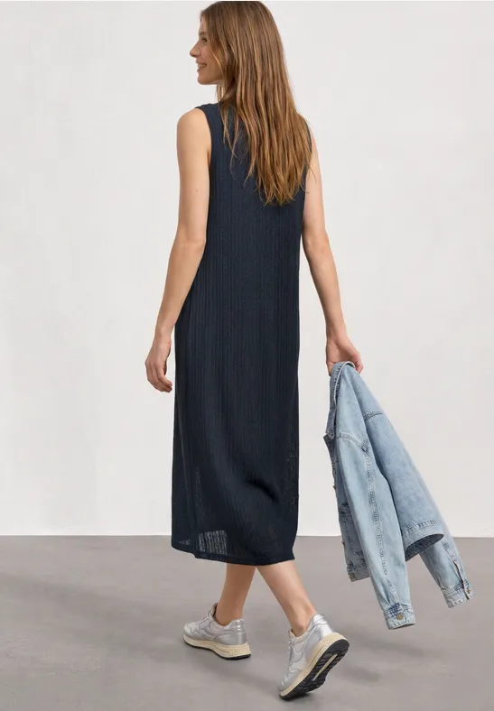 Vrouw van achteren in zwarte mouwloze midijurk, draagt een spijkerjasje en witte sneakers.