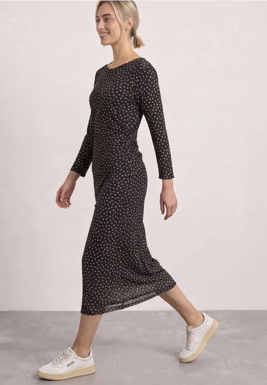 Une femme souriante marche en robe midi noire à pois blancs et baskets beiges.