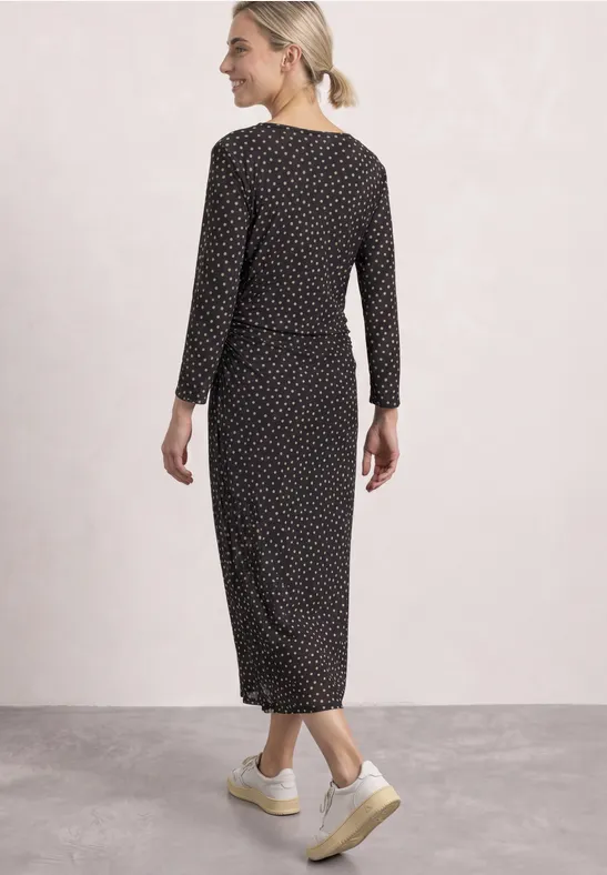 Femme de dos en robe midi noire à pois blancs à manches longues, posée sur un sol neutre.