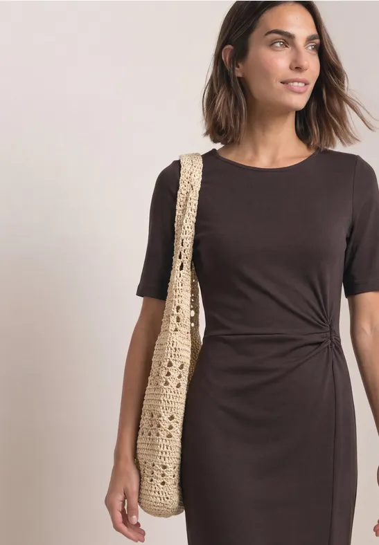 Femme en robe marron mi-longue avec noeud latéral et manches courtes, portant un sac en crochet.