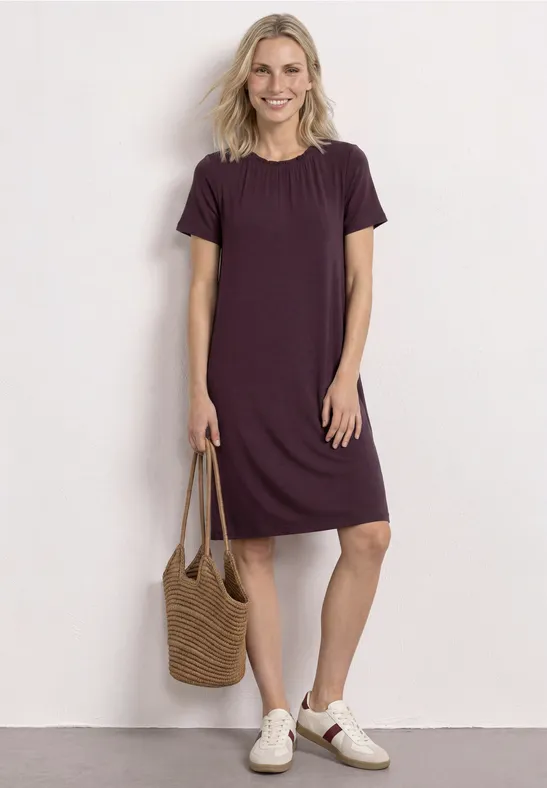 Une femme porte une robe t-shirt violette jusqu'aux genoux, un sac en paille tressé et des baskets blanches.