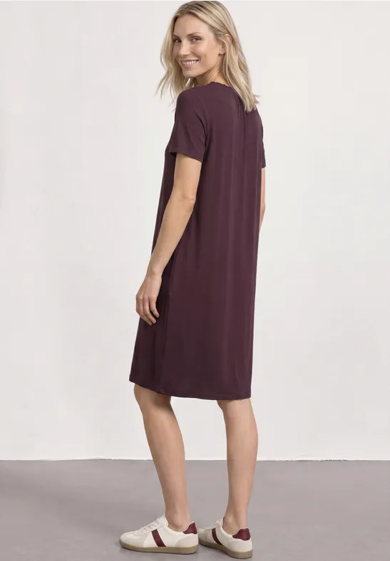 Une femme de profil porte une robe-t-shirt violette et des baskets blanches.