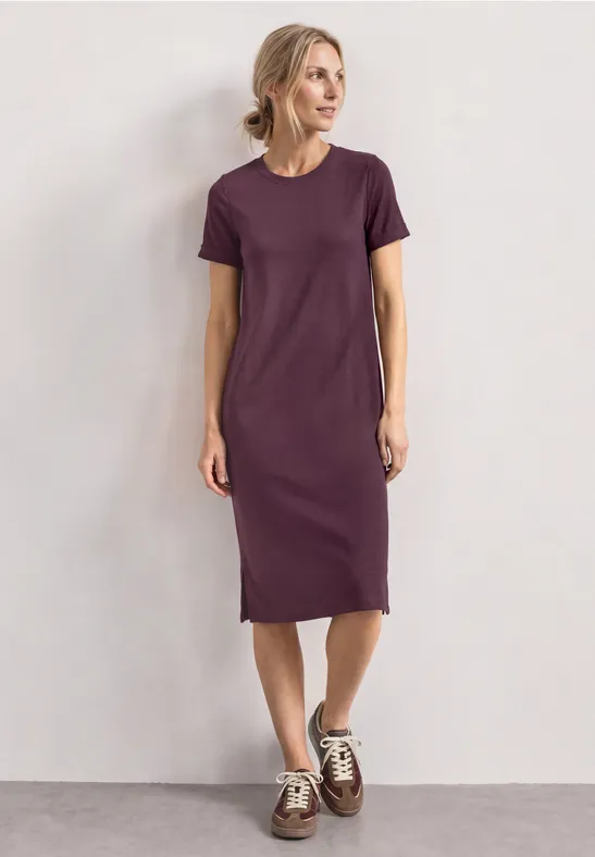 Vrouw in eenvoudig paarse midi T-shirtjurk met witte sneakers voor een neutrale achtergrond.