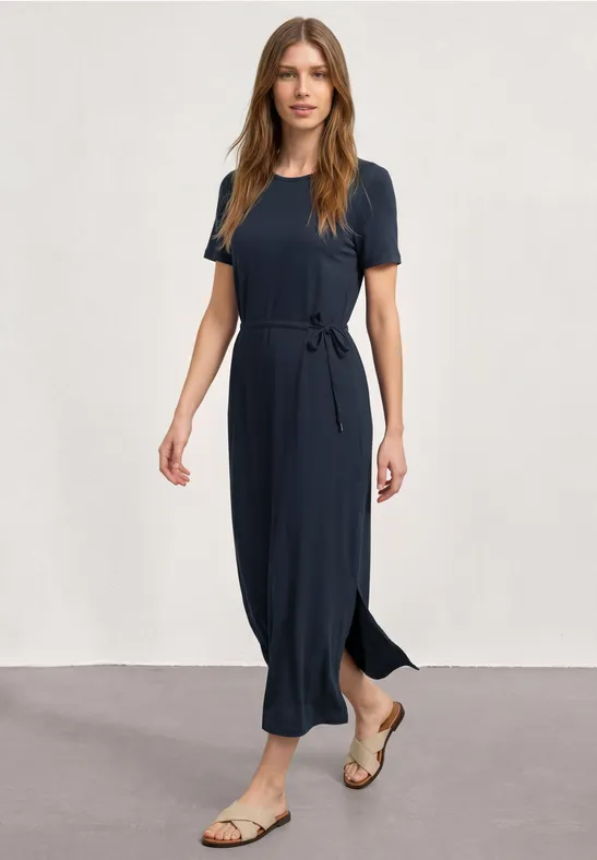 Femme en robe mi-longue bleu marine avec ceinture nouée et sandales sur fond neutre.