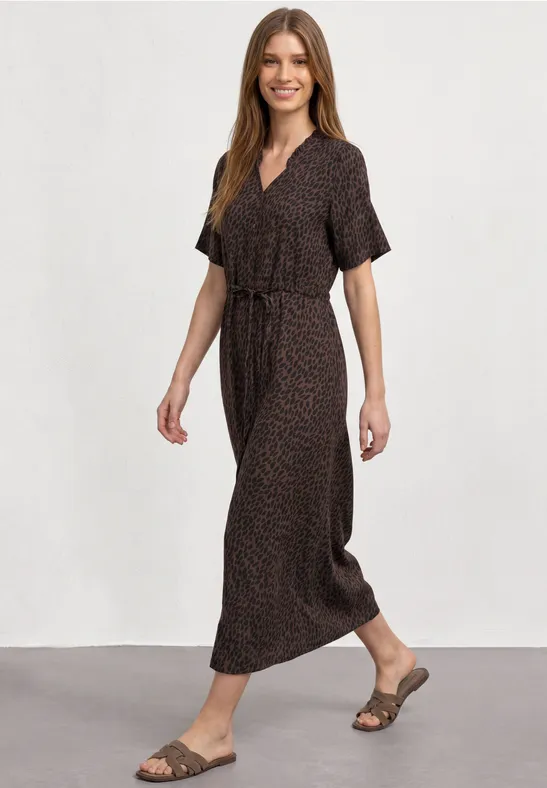 Frau in braunem Maxikleid mit kurzem Arm, Bindegürtel und Sandalen, die lächelnd geht.