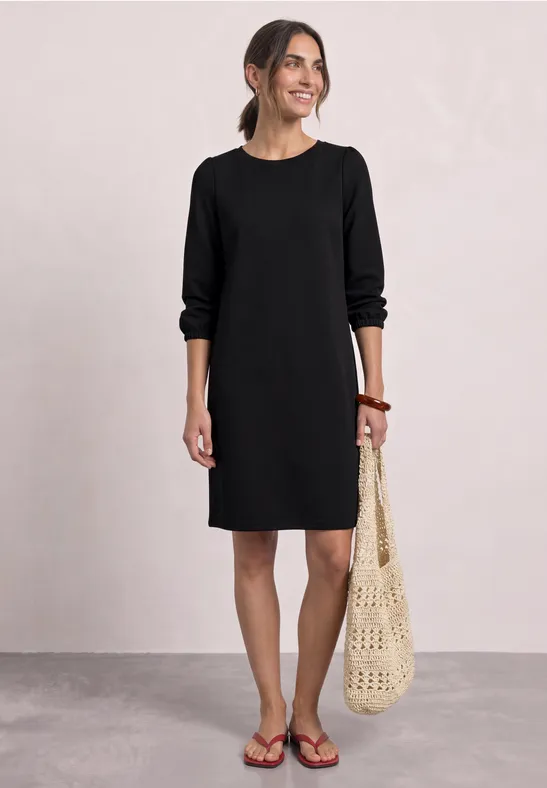 Frau im schwarzen, knielangen Kleid mit langen Ärmeln und gehäkelter Tasche, lächelnd und in Sandalen.