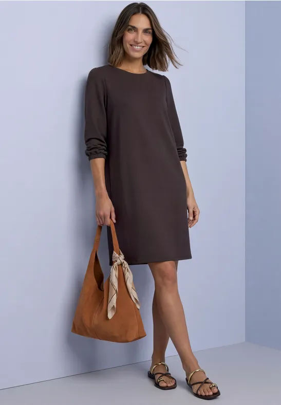 Frau in lockerem, braunem Kleid mit lässiger Handtasche und Sandalen vor pastellfarbener Wand.
