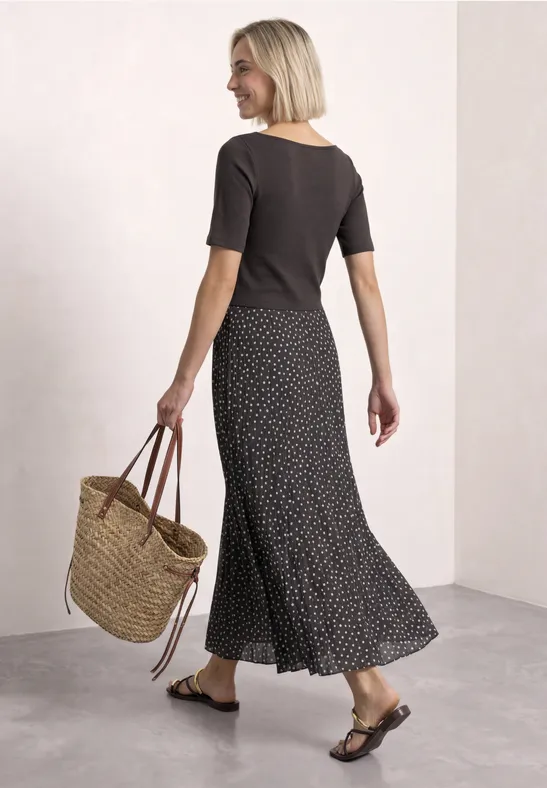 Femme en top noir, jupe longue à pois et sac en osier, pose souriante de profil.