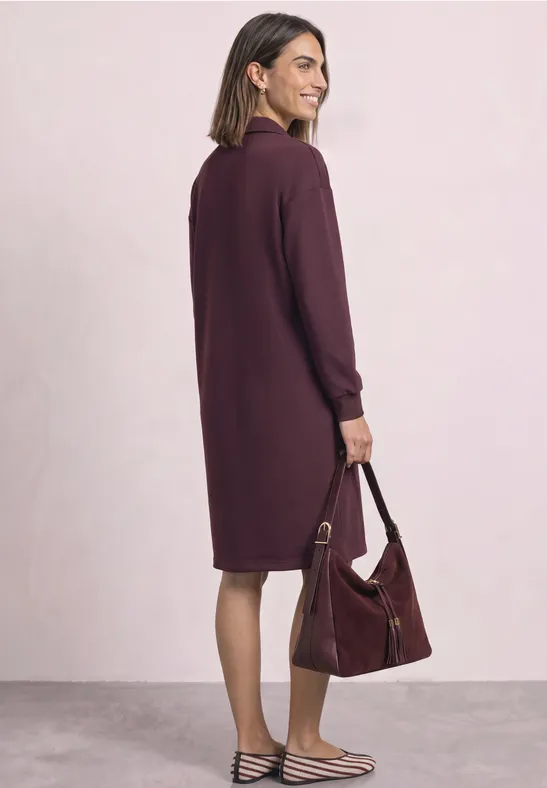 Eine Frau trägt ein knielanges, locker sitzendes, dunkelviolettes Kleid und hält eine passende Handtasche.
