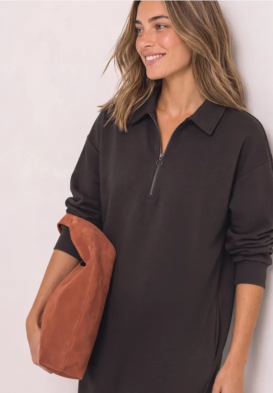 Frau in schwarzem, langärmligem Kleid mit halbem Reißverschluss, lächelnd, mit großer brauner Tasche.