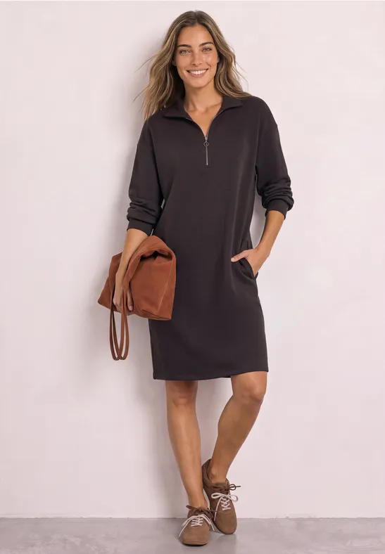 Frau in schwarzem Sweatkleid mit halbem Reißverschluss, Taschen und braunen Accessoires.