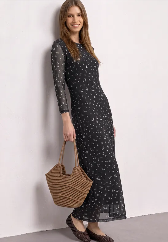 Eine Frau trägt ein langes schwarzes Kleid mit floralem Muster und hält eine Korbtasche.