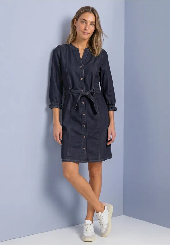 Une femme porte une robe-chemise en jean foncé ceinturée et des baskets blanches, debout devant un mur clair.