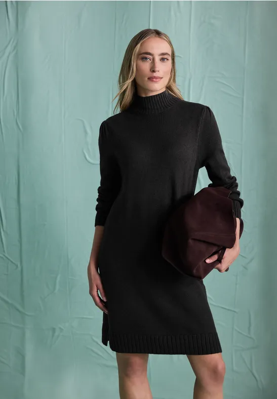 Frau in schwarzem Strickkleid und Rollkragen, hält eine bordeauxrote Tasche vor grünem Hintergrund.