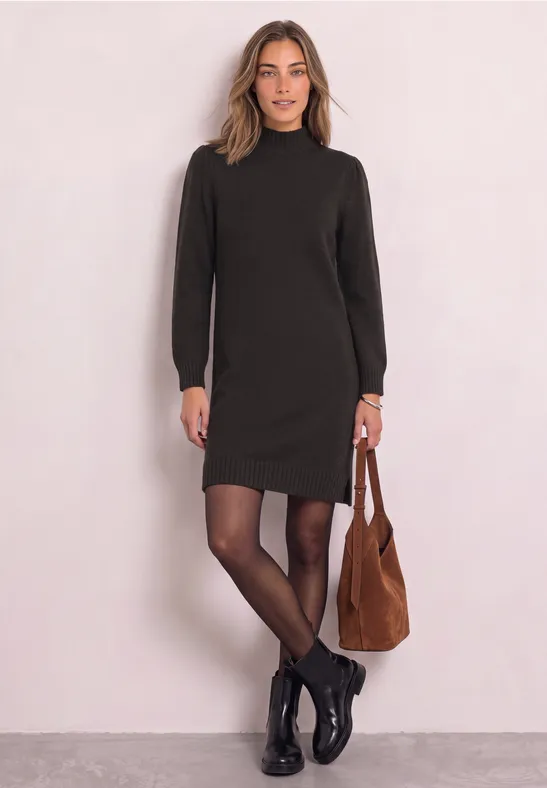 Frau trägt dunkles, langärmeliges Kleid, schwarze Strumpfhose, Boots und braune Schultertasche vor hellem Hintergrund.